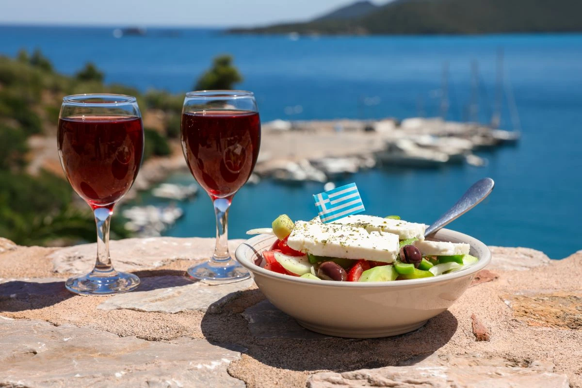 A Greek salad and two glasses of wine with a Greek flag and a peaceful harbour in the background